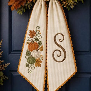 May include: A cream-colored hanging fabric decoration with vertical stripes and a scalloped orange border. One side features an embroidered design of orange, white, and green pumpkins with autumn leaves. The other side displays a large brown letter S.