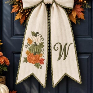 May include: A cream-colored fabric bow with olive green scalloped edges. The left tail features an embroidered stack of three pumpkins in white, green, and orange with autumn leaves. The right tail displays a large, embroidered olive green letter W.