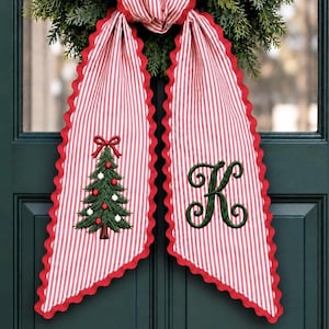 May include: A decorative fabric bow with red and white vertical stripes and scalloped edges. The left tail features an embroidered green Christmas tree with red and white ornaments, while the right tail displays a large, dark green letter K.