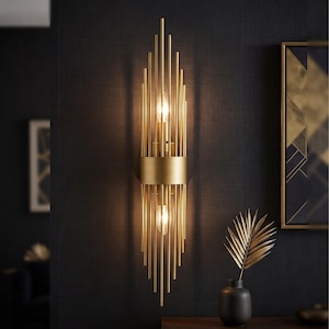 May include: A vertical wall sconce featuring a central gold-toned cylindrical band surrounded by numerous thin, parallel gold metal rods of varying lengths that extend upward and downward, housing two clear light bulbs emitting a warm glow.