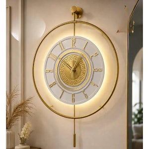 May include: A circular wall clock with a gold metal frame and a white face featuring gold Roman numerals. The center has a gold disc with a Greek key pattern and gold hands. A vertical gold rod supports the clock and a cylindrical pendulum.