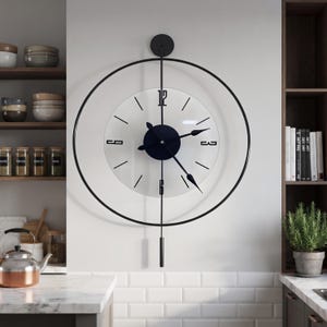 Op de afbeelding: Een wandklok met een grote zwarte cirkelvormige metalen rand rondom een heldere glazen wijzerplaat. De klok heeft zwarte wijzers, een zwarte centrale naaf en zwarte cijfers op de posities 12, 3, 6 en 9. Een verticale stang loopt naar een slinger.