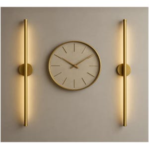 May include: A circular wall clock with a light beige face and thin gold hands, flanked by two vertical gold wall sconces. Each sconce features a long, slender cylindrical rod with integrated warm lighting that illuminates the wall surface.