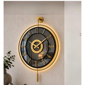 May include: A round wall clock featuring a black glass face with gold-colored numerals and hands. The clock is framed in a gold-colored metal ring with integrated backlighting and includes a gold-colored pendulum and a top mounting bracket.
