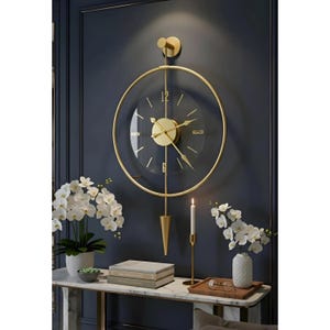 Op de afbeelding: Een ronde wandklok met een goudkleurig metalen frame en een transparante wijzerplaat. De klok heeft gouden wijzers, gouden uurmarkeringen en een verticale gouden staaf die uitloopt in een puntig gouden slingergewicht aan de onderzijde.