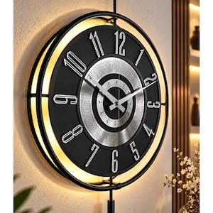Puede incluir: Un reloj de pared circular con marco negro y esfera negra que presenta números plateados y un anillo central plateado con un patrón griego. El reloj incluye una varilla de montaje vertical, un péndulo y retroiluminación cálida integrada.
