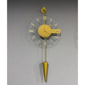 Peut inclure: Une horloge murale dorée dotée d'un cadran circulaire transparent avec des chiffres dorés aux positions 12 et 6. L'horloge présente un cadran central doré, des aiguilles dorées et une longue tige verticale dorée avec un poids de balancier pointu.