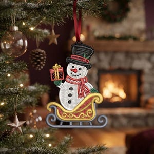 May include: An embroidered snowman ornament wearing a black top hat and a red scarf, holding a small gift box while sitting in a gold and gray sleigh. The ornament is suspended by a red ribbon and features detailed stitching throughout the design.