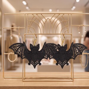 May include: A pair of black bat-shaped earrings featuring intricate lace-like wing details and gold-toned leverback hooks. The earrings are displayed on a gold metal jewelry stand, which also holds several delicate bracelets and necklaces.