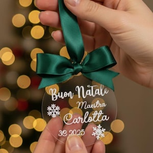 Può includere: Una decorazione natalizia rotonda in acrilico trasparente con un fiocco in nastro di raso verde scuro sulla parte superiore. L'ornamento presenta la scritta bianca Buon Natale, Maestra, Carlotta e 3e 2025, con due grafiche di fiocchi di neve.