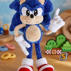 May include: A plush, crocheted Sonic the Hedgehog figure featuring a vibrant blue body, tan muzzle and belly, white gloves, and red sneakers with white cuffs and grey soles. The character has large black eyes and is posed with one finger raised.