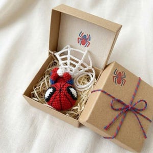 May include: A small, red and blue crocheted superhero figure with large white eyes, accompanied by a white crocheted web, nestled in a brown cardboard gift box. The box lid features a red and blue spider icon and is tied with matching twine.