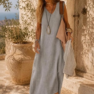 Pode incluir: Um vestido midi sem mangas cinza-claro com decote em V, combinado com um chapéu de palha com faixa preta, um colar longo com pingente, uma bolsa de ombro bege, um acessório de tecido cinza-claro e sandálias castanhas de tiras com sola plana.