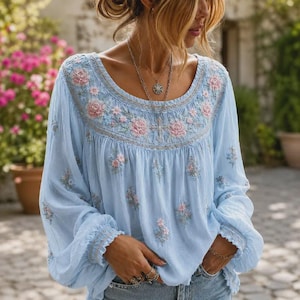 Op de afbeelding: Een lichtblauwe blouse met lange mouwen, een ronde halslijn en gedetailleerd bloemenborduursel in roze en lichtblauwe tinten. Het kledingstuk heeft geplooide mouwboorden en een losvallend, vloeiend silhouet, aangevuld met een zilveren kruishanger.
