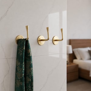 Peut inclure: Trois patères murales en or brossé avec une base circulaire et un bras incurvé pointant vers le haut. Une écharpe vert foncé à motif floral doré est suspendue au crochet le plus à gauche, illustrant le design fonctionnel de ces fixations.