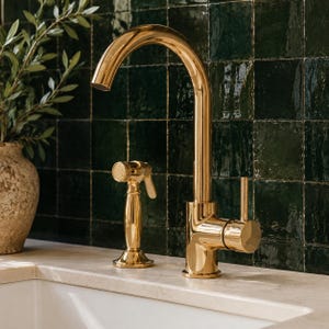 May include: A polished gold kitchen faucet with a high-arc spout and a matching side sprayer. The faucet features a single lever handle on the right side, providing a sleek, metallic finish that contrasts with the dark green tiled wall behind it.