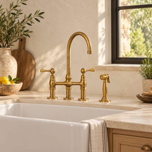 May include: A bridge-style kitchen tap with a high-arc spout and two lever handles in a brushed brass finish. The tap includes a matching side sprayer positioned to the right, all mounted on a light-coloured countertop above a white sink.