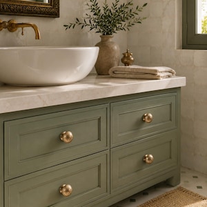 Puede incluir: Un mueble de baño verde salvia con cuatro cajones y pomos redondos de latón texturizado. El mueble tiene una encimera de piedra blanca, un lavabo de cerámica blanca y un grifo de latón. Una toalla de color beige descansa sobre la encimera.