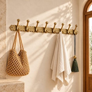 May include: A long, horizontal brass wall rack featuring eight upward-curving hooks and an intricate, embossed floral pattern along the base. A tan woven net tote bag, a plain white cloth, and a dark green decorative tassel hang from the hooks.