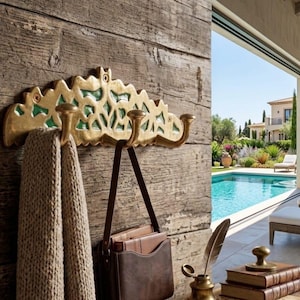 May include: A gold-toned wall-mounted coat rack featuring three hooks and an ornate, arched top section with green accents. The rack is attached to a textured wooden surface, with a beige knit towel and a brown leather bag hanging from the hooks.