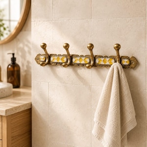 Puede incluir: Un perchero de pared con cuatro ganchos de tono latón y una barra horizontal decorativa con azulejos incrustados de patrón de diamantes amarillos y blancos. Una toalla beige cuelga del gancho derecho de este accesorio metálico.