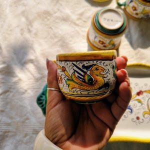 Puede incluir: Una taza de cerámica con un motivo de dragón alado pintado a mano en amarillo, verde y azul sobre un fondo blanco. La taza tiene un borde y una base amarillos, con otras tazas a juego y una bandeja rectangular visibles al fondo.