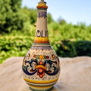 Puede incluir: Una aceitera de cerámica con cuello estrecho y base bulbosa, decorada con motivos florales pintados a mano en azul, verde, naranja y amarillo. La palabra OLIO está impresa en letras negras en el cuello y tiene un vertedor de metal arriba.
