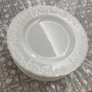 Può includere: Una pila di piatti in ceramica bianca con un centro circolare liscio e un bordo largo e smerlato decorato con un motivo a foglie in rilievo. I piatti hanno una finitura lucida che riflette la luce su tutta la superficie.
