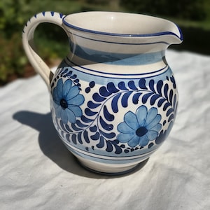 Può includere: Una brocca in ceramica con base color crema, decorata con motivi floreali e foglie blu dipinti a mano. Il recipiente presenta un manico curvo, un ampio beccuccio per versare e fasce blu orizzontali attorno al collo e alla base.