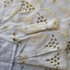May include: A white fabric textile featuring intricate eyelet embroidery with pale yellow thread detailing. The edges are scalloped with a decorative lattice pattern and floral stitching, showcasing a delicate needlework design on the cloth.