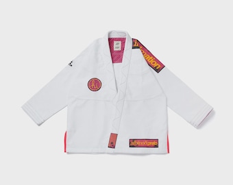 Könnte beinhalten: Eine weiße Kampfkunst-Gi-Jacke mit langen Ärmeln, einem dicken Kragen mit Wellennähten und einem rosa-gelb gestreiften Innenfutter. Sie verfügt über einen runden Aufnäher auf der Brust, einen rechteckigen Aufnäher mit dem Text albino&preto und einen Schulteraufnäher mit dem Text in motion.