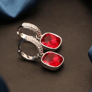 May include: A pair of silver-toned hoop earrings featuring a hinged closure and a row of small clear stones along the front. Each earring suspends a square-cut, vibrant red gemstone set in a polished silver-toned frame, creating a classic look.
