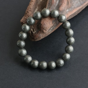 May include: A circular bracelet composed of numerous spherical, metallic gray pyrite beads strung together. The beads feature a textured, reflective surface with a natural luster, arranged in a continuous loop to form a simple, unadorned jewelry piece.
