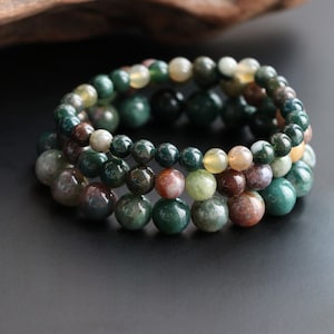May include: A beaded bracelet composed of polished spherical stones in various shades of deep forest green, moss green, earthy brown, and translucent cream. The beads are strung together in a circular formation, showcasing natural color variations.