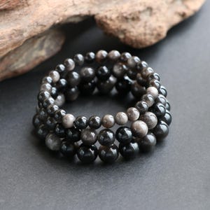 May include: A stack of three beaded bracelets featuring polished, spherical beads in shades of black, charcoal, and silver-grey. The beads exhibit a smooth, reflective surface with subtle metallic sheen, arranged in a circular, elasticised formation.
