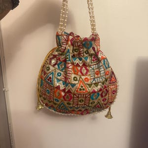 以下が含まれることがあります： クリーム色のベースに鮮やかなマルチカラーの幾何学模様の刺繍と小さなスパンコールが施された巾着バッグ。パールビーズのハンドル2本と、引き紐の付け根にゴールドトーンの金属製タッセルが付いています。