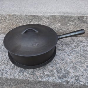 May include: A black cast iron skillet with a matching lid and a long handle. The lid features a small central handle and a circular embossed logo with the letters O&S. The cookware has a matte finish and a deep, cylindrical shape for cooking.