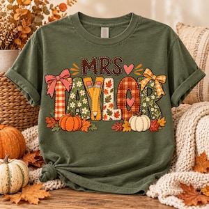 May include: An olive green t-shirt featuring the text MRS TAYLOR in a decorative font. The letters are filled with patterns including red plaid, autumn leaves, and daisies. A yellow pencil forms the letter Y, and orange and white pumpkins are below.