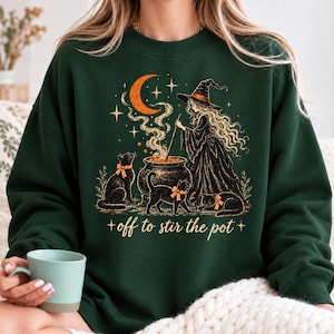 May include: A dark green crewneck sweatshirt featuring a graphic of a witch stirring a cauldron with three black cats wearing orange plaid bows. Above the scene is an orange crescent moon and stars. The text reads, off to stir the pot.