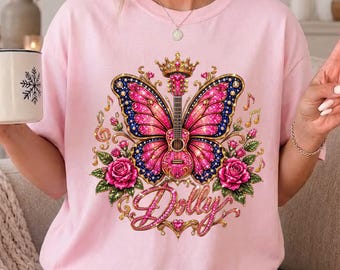 May include: A light pink t-shirt featuring a graphic print of a guitar with butterfly wings, a crown, and roses. The design includes musical notes and the word Dolly in cursive script. The shirt has a crew neckline and short sleeves.