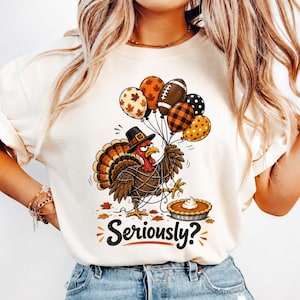 Könnte beinhalten: Ein cremefarbenes T-Shirt mit einer Grafik eines Truthahns mit schwarzem Hut, der sich in Schnüren von fünf unterschiedlich gemusterten Luftballons verheddert hat und neben einem Kürbiskuchen steht, mit dem schwarzen Schriftzug Seriously? darunter.