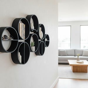May include: A wall-mounted shelving unit composed of multiple interconnected black circular frames of varying sizes. The shelves hold various items including books, a small potted plant, a white vase, and a pair of sunglasses on the flat surfaces.