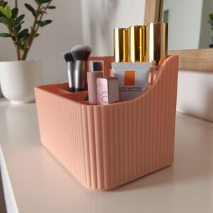 May include: A peach-coloured plastic desk organiser with a vertical ribbed texture and multiple compartments. It contains various cosmetic items, including makeup brushes, small tubes, and a La Roche-Posay skincare product with a white and orange label.