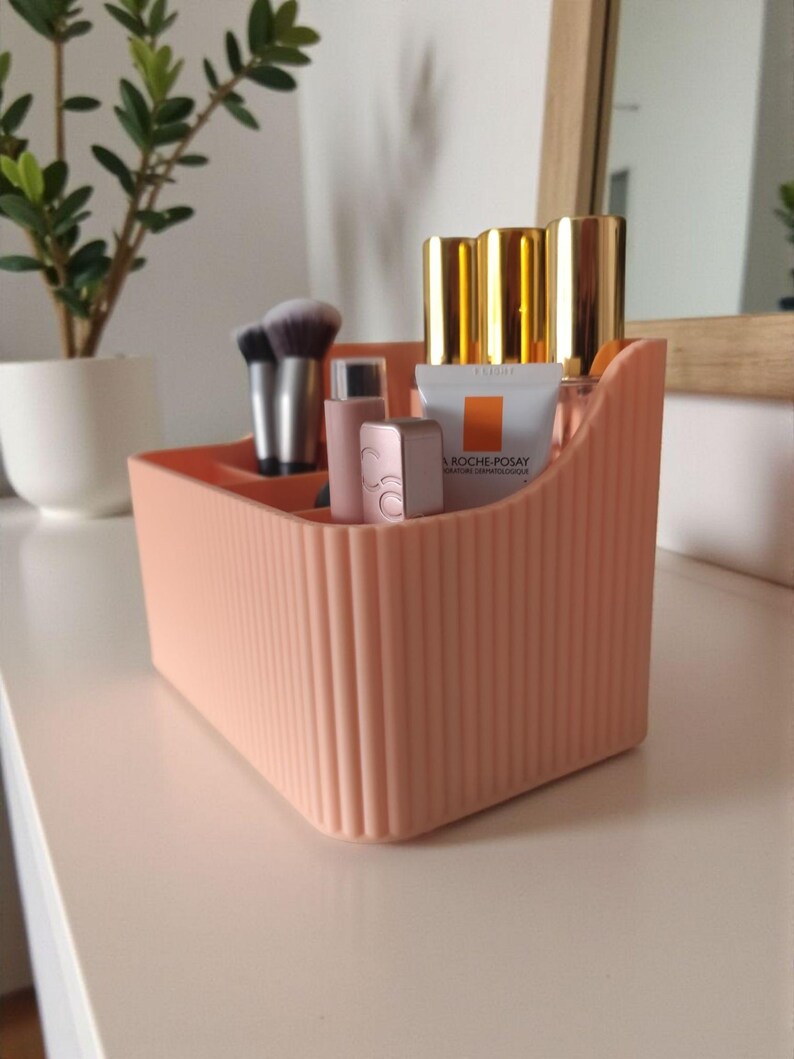 May include: A peach-coloured plastic desk organiser with a vertical ribbed texture and multiple compartments. It contains various cosmetic items, including makeup brushes, small tubes, and a La Roche-Posay skincare product with a white and orange label.