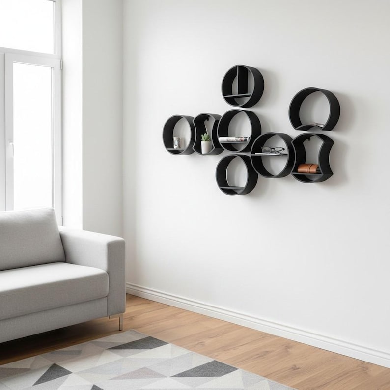 May include: A collection of black wall-mounted shelves in various circular and geometric shapes arranged in a cluster. The shelves feature flat horizontal surfaces for storage and display, creating a modular storage system on a plain white wall.