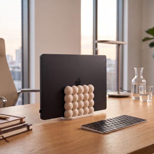 May include: A matte beige vertical laptop stand featuring a grid of rounded, bubble-like spheres on the front. The stand holds a black laptop upright, providing a stable base for the device on a wooden desk surface alongside a keyboard and notebooks.
