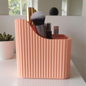 May include: A peach-coloured plastic desk organiser with a vertical ribbed texture and a curved top edge. The container holds various items including a makeup brush with black and white bristles, a gold-capped tube, and a small spray bottle.