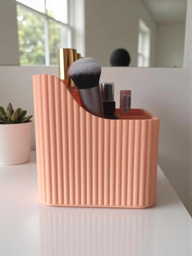May include: A peach-coloured plastic desk organiser with a vertical ribbed texture and a curved top edge. The container holds various items including a makeup brush with black and white bristles, a gold-capped tube, and a small spray bottle.