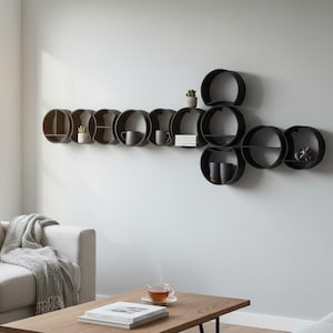 May include: A wall-mounted shelving unit composed of multiple interconnected black circular frames of varying sizes. The shelves hold small potted plants, ceramic mugs, and a stack of books, creating a modular storage display on the wall.
