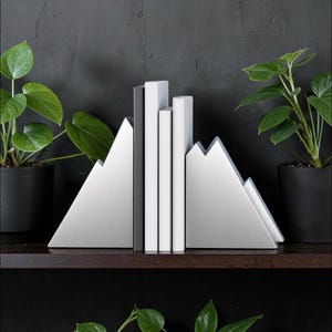 Peut inclure: Une paire de serre-livres en m&eacute;tal argent&eacute; en forme de sommets de montagne, maintenant trois livres &agrave; la verticale. Les serre-livres ont une finition bross&eacute;e et une silhouette triangulaire, plac&eacute;s entre deux plantes vertes en pot sur une &eacute;tag&egrave;re sombre.